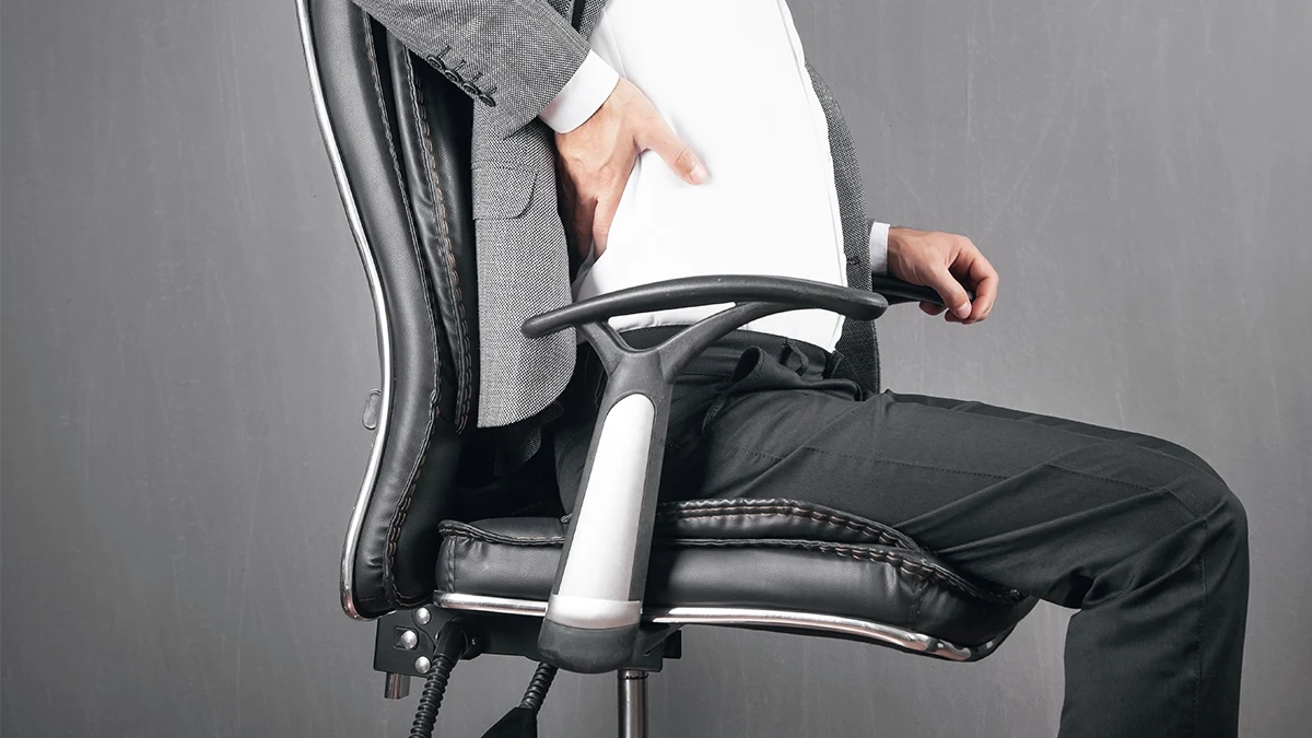 Homem sentado em cadeira de escritório ergonômica preta, com expressão de desconforto devido a dores nas costas em uma sala de escritório.